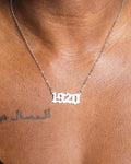 Silver '1920' necklace on a person's neck with visible skin and tattoo.