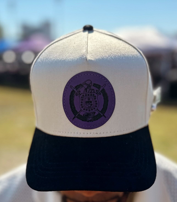 A cream and black snapback cap with a purple circular patch with the shield of Omega Psi Phi on the front, shown on a person outdoors. 