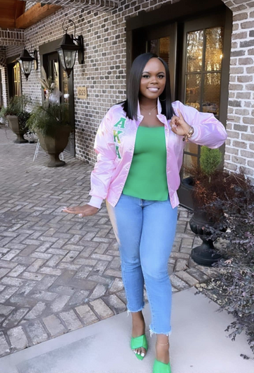 A woman stands outdoors in front of a brick building, wearing a light pink bomber jacket with green and white greek lettering AKA, a bright green top, light blue jeans, and green high heels.
