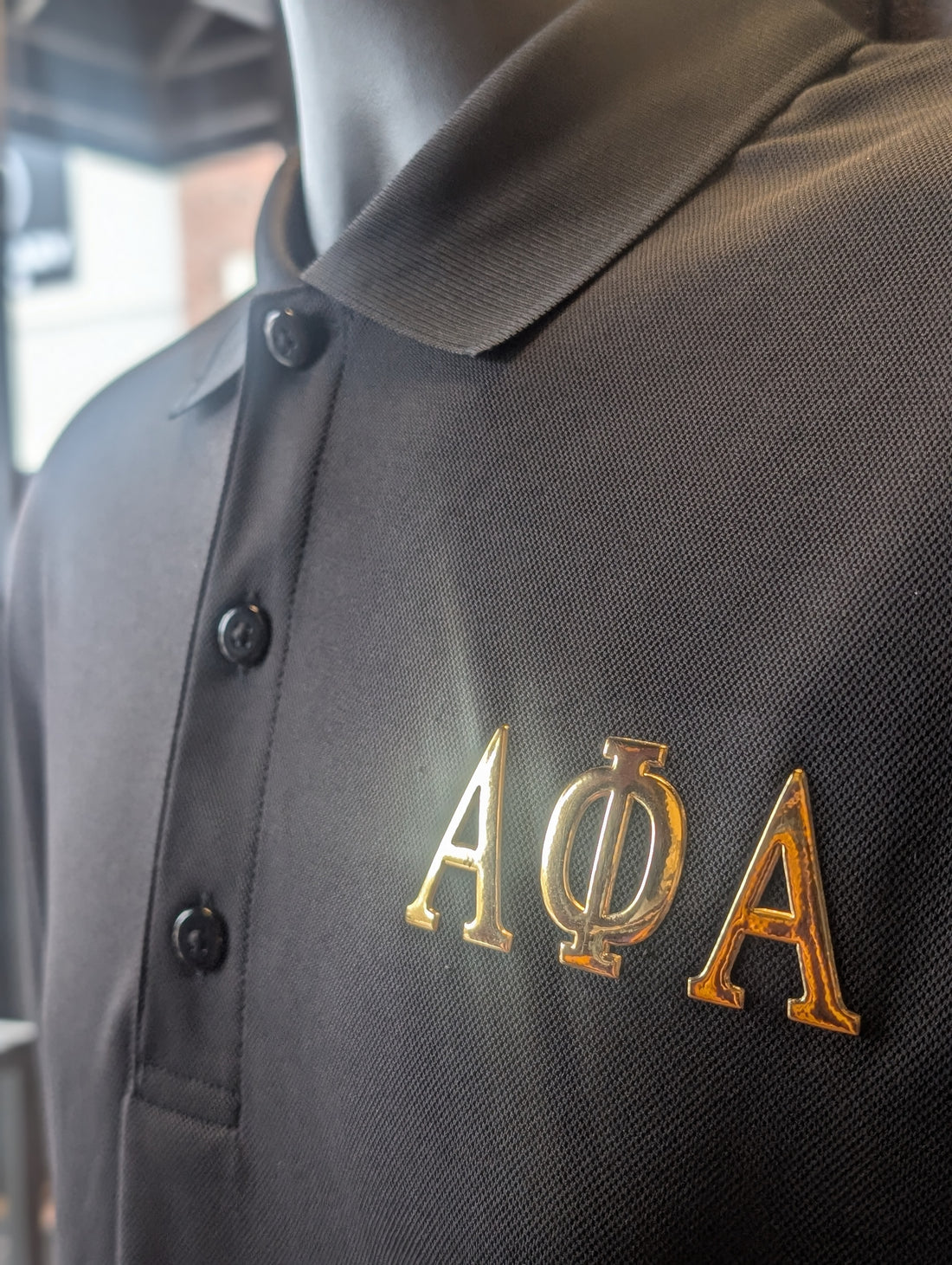 Black polo shirt with gold greek letters for alpha on the left chest
