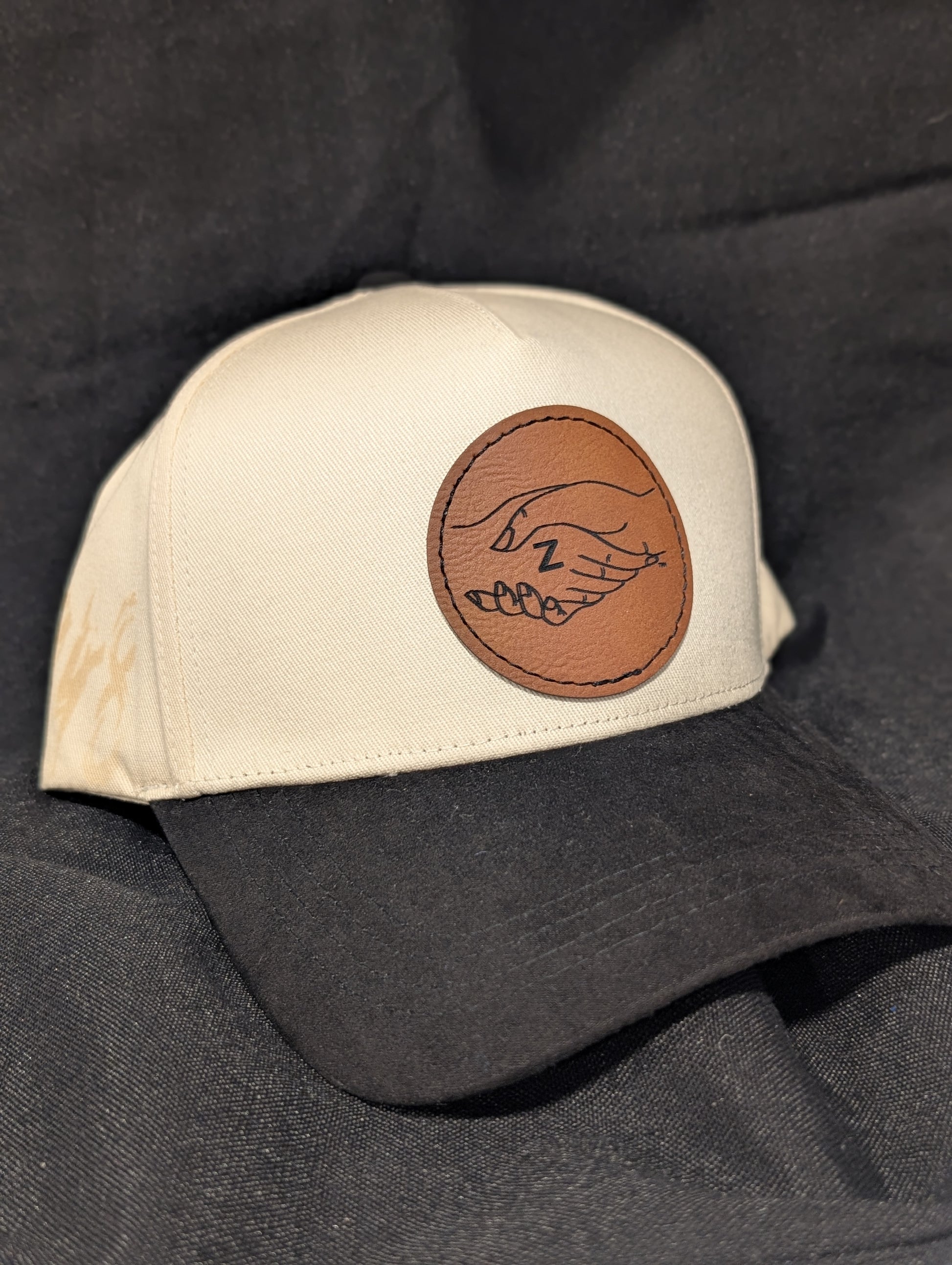 A baseball cap with an off-white crown and a black curved brim, featuring a circular brown faux-leather patch on the front with the zeta amicae logo laser engraved on it
