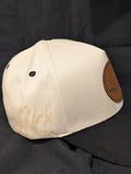 A baseball cap with an off-white crown and a black curved brim, featuring a circular brown faux-leather patch on the front with the zeta amicae logo laser engraved on it