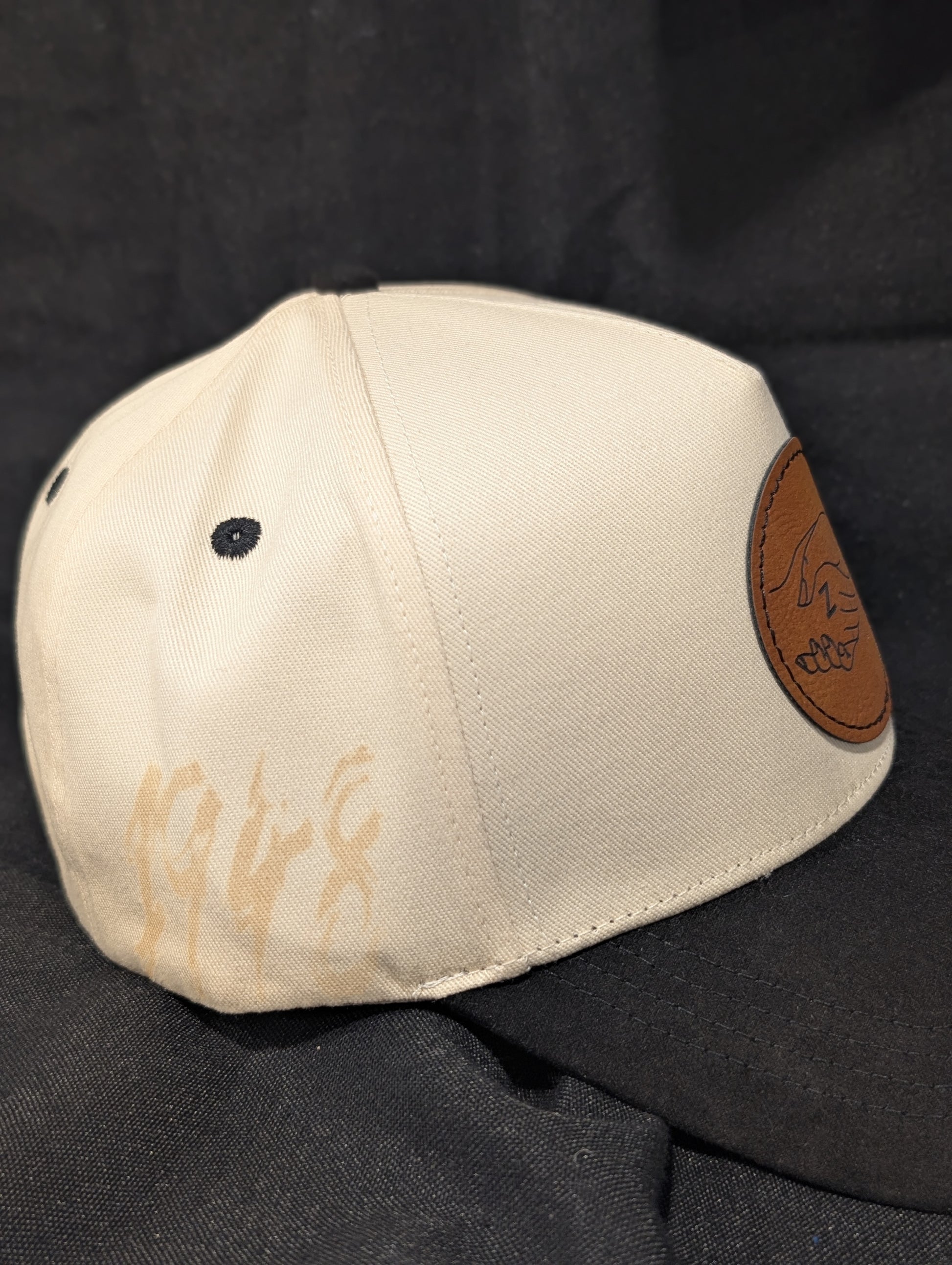 A baseball cap with an off-white crown and a black curved brim, featuring a circular brown faux-leather patch on the front with the zeta amicae logo laser engraved on it