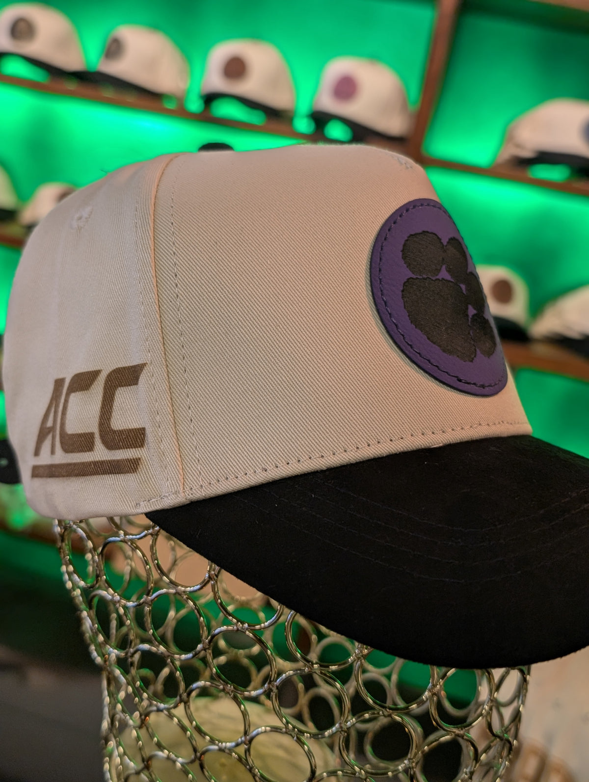 Beige cap with black brim and purple ACC logo on a green background