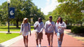 Four students walking on a college campus path with trees and a flag in the background.
