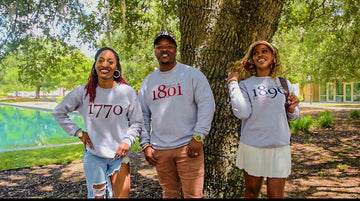 Three people wearing collegiate sweatshirts, standing by a tree in a park-like setting.