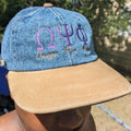 Blue denim cap with embroidered symbols and text, worn by a person outdoors.