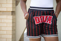 Black shorts with red pinstripes and 'DIVA' text worn by a person, against a beige brick wall.
