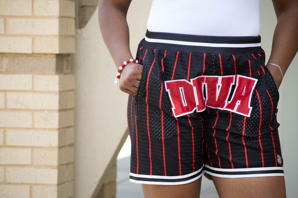 Black shorts with red pinstripes and 'DIVA' text worn by a person, against a beige brick wall.
