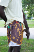 A person outdoors wearing patterned athletic shorts with a brown base, gold lettering, and white mesh lining, paired with a light-colored t-shirt.