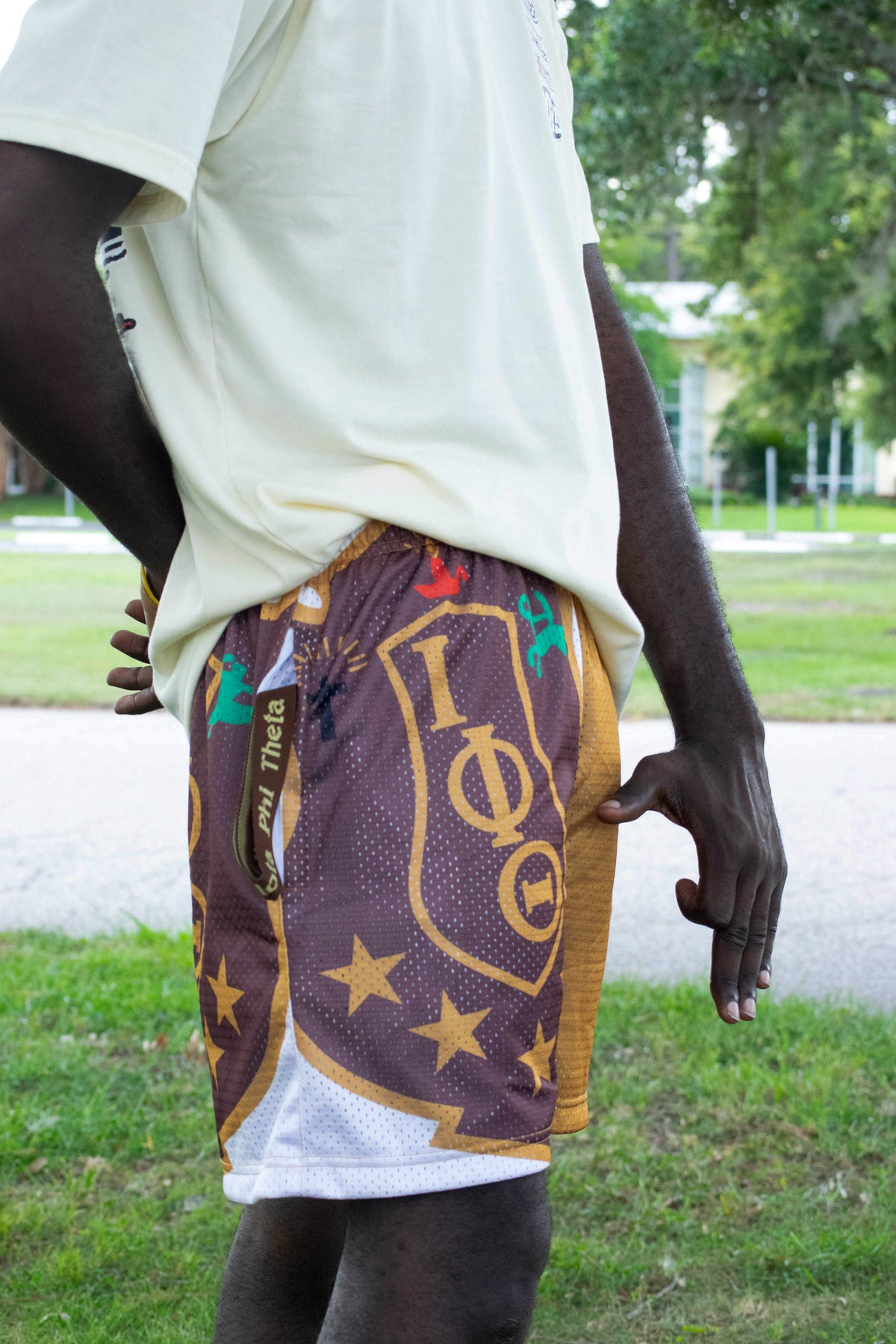 A person outdoors wearing patterned athletic shorts with a brown base, gold lettering, and white mesh lining, paired with a light-colored t-shirt.