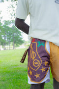 A person outdoors wearing athletic shorts with a purple mesh front panel featuring gold Greek letters and stars, a white mesh side panel, and a gold side flap, paired with a cream t-shirt.