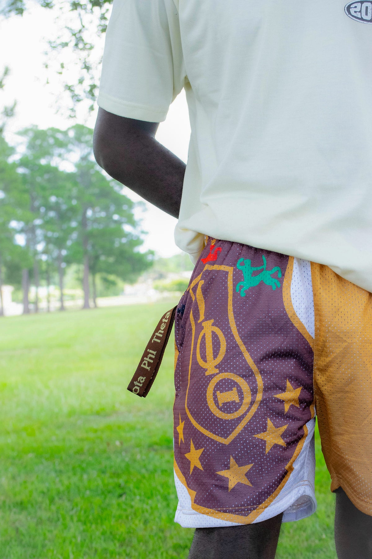 A person outdoors wearing athletic shorts with a purple mesh front panel featuring gold Greek letters and stars, a white mesh side panel, and a gold side flap, paired with a cream t-shirt.