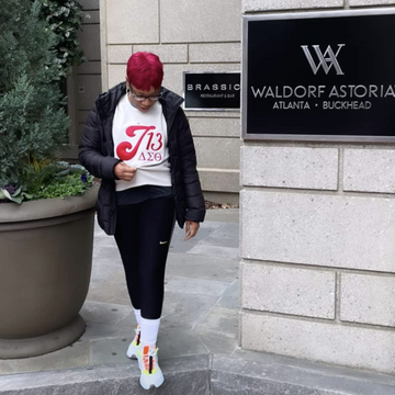 A Delta walking outdoors next to a stone building wall with a black Waldorf Astoria sign, wearing a black puffer jacket, cream crewneck with a red J13 graphic, black leggings, white socks, and colorful sneakers.