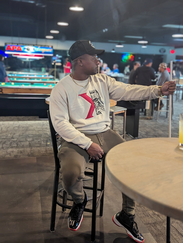 Man sitting wearing a cream sweatshirt with a red logo and black cap.
