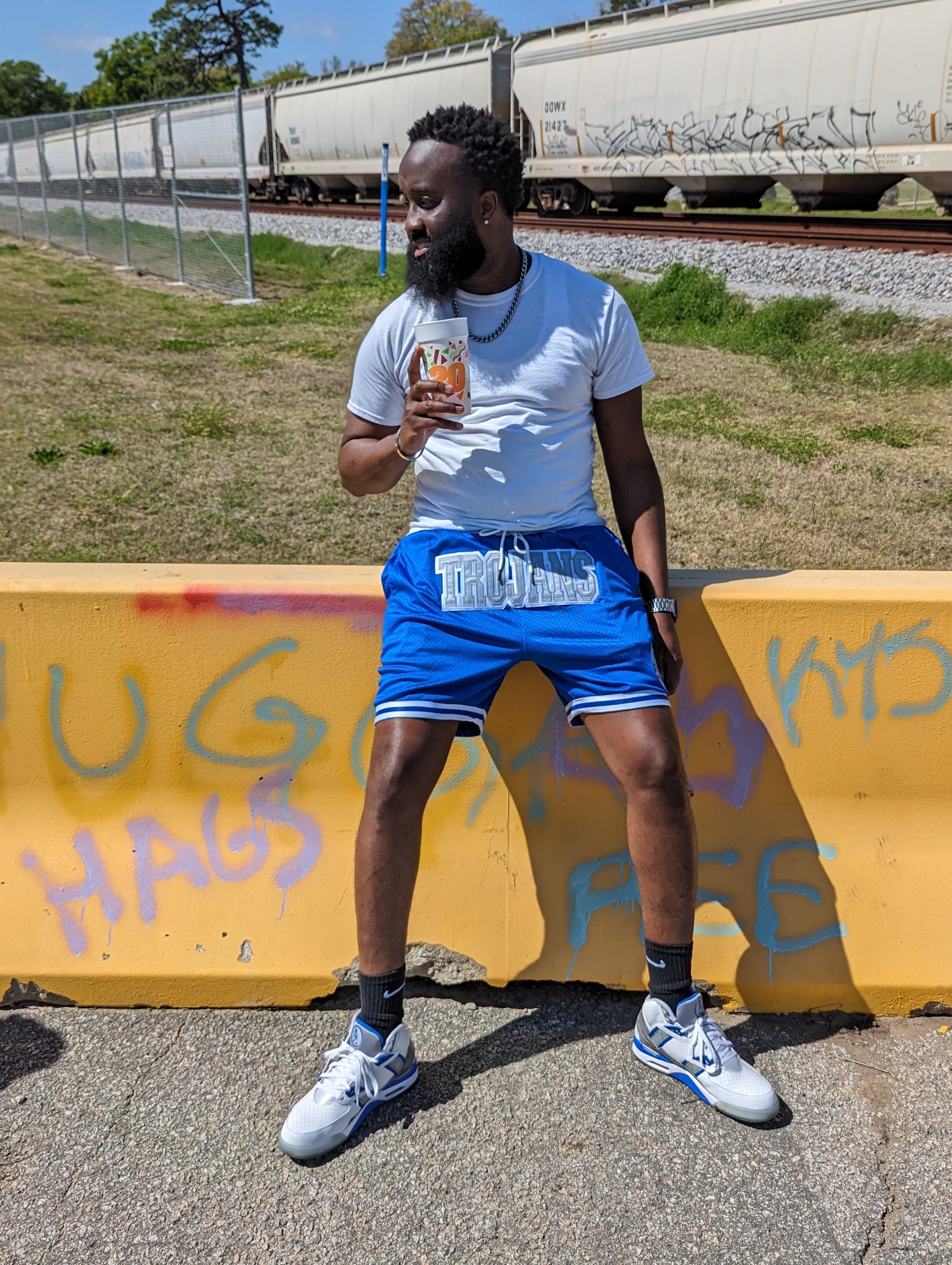 Man holding a drink against a graffiti-covered wall with train tracks in the background