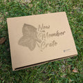 A cardboard gift box with the text 'NEW MEMBER CRATE' printed on it, placed on grass with an ivy leaf design beside it.