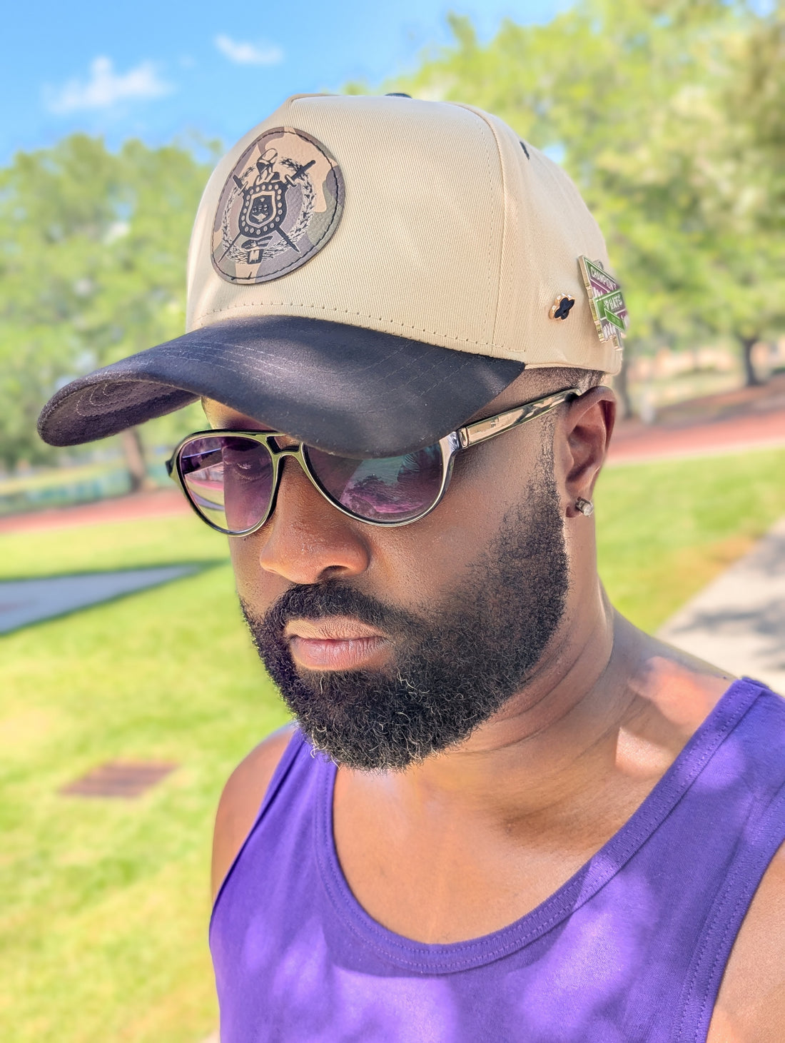 Man wearing a beige cap with an omega psi phi crest and sunglasses, standing outdoors with greenery in the background