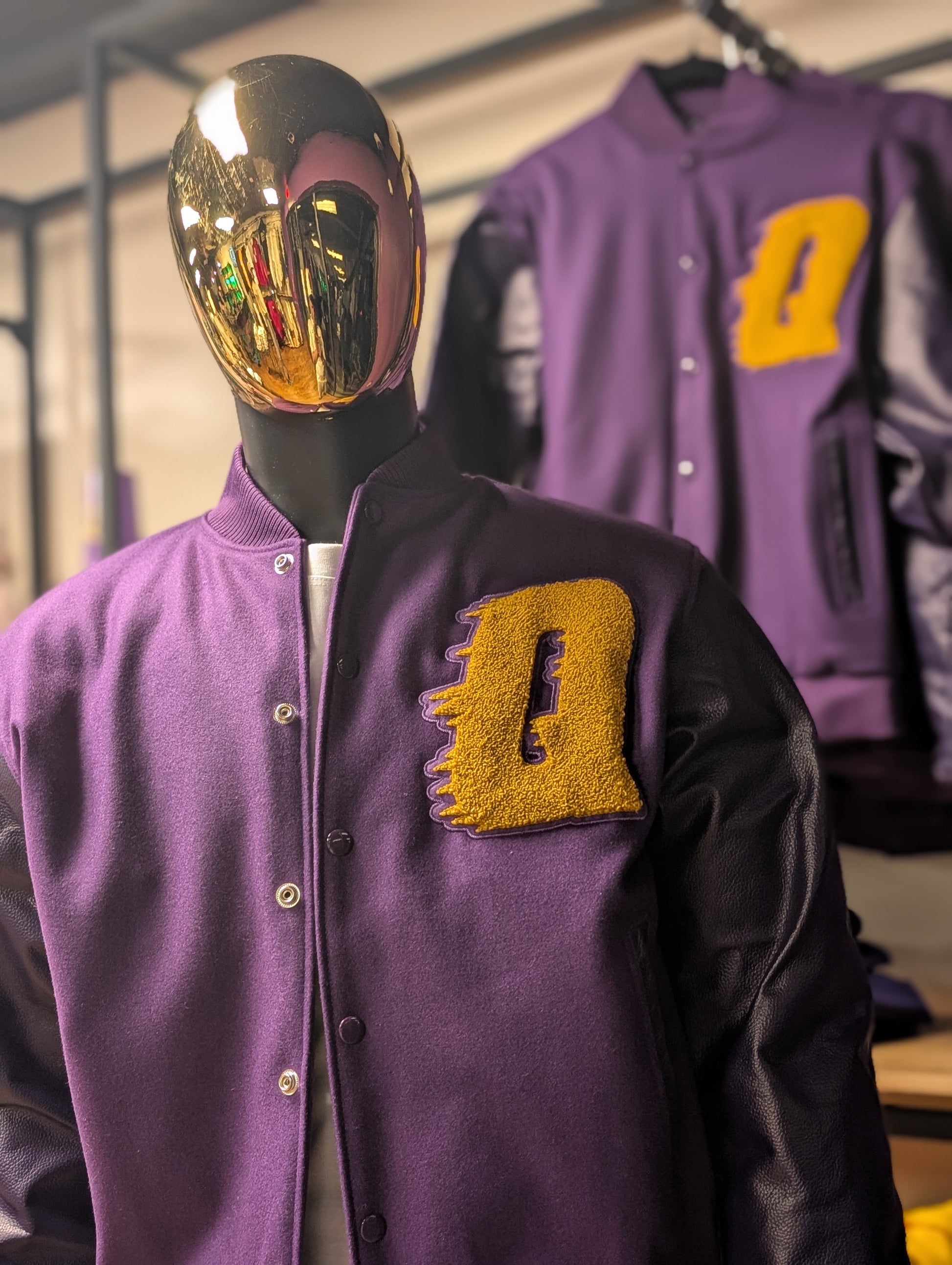 A purple varsity jacket with a gold Omega Psi Phi logo on the front, displayed on a mannequin with a golden mask.