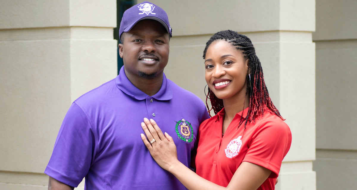 Husband & wife wearing purple and red polo shirts with greek org shields, standing in front of a beige building.
