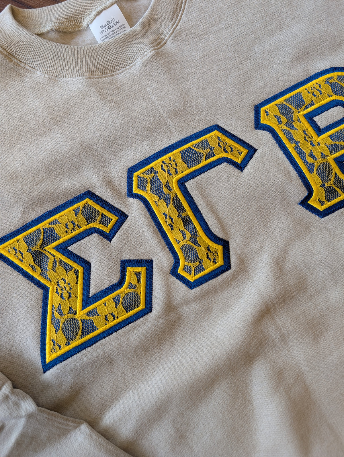 Close-up of a beige crewneck sweatshirt with blue and yellow Greek letters 'SGR' embroidered on the front.