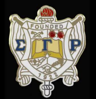 A close-up of a crest-style emblem or pin on a black background, featuring Greek letters, an open book, crossed swords, small red rose motifs, and the text 'FOUNDED 1922'.