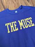 purple t-shirt with 'THE MUSE' text in yellow on a wooden surface