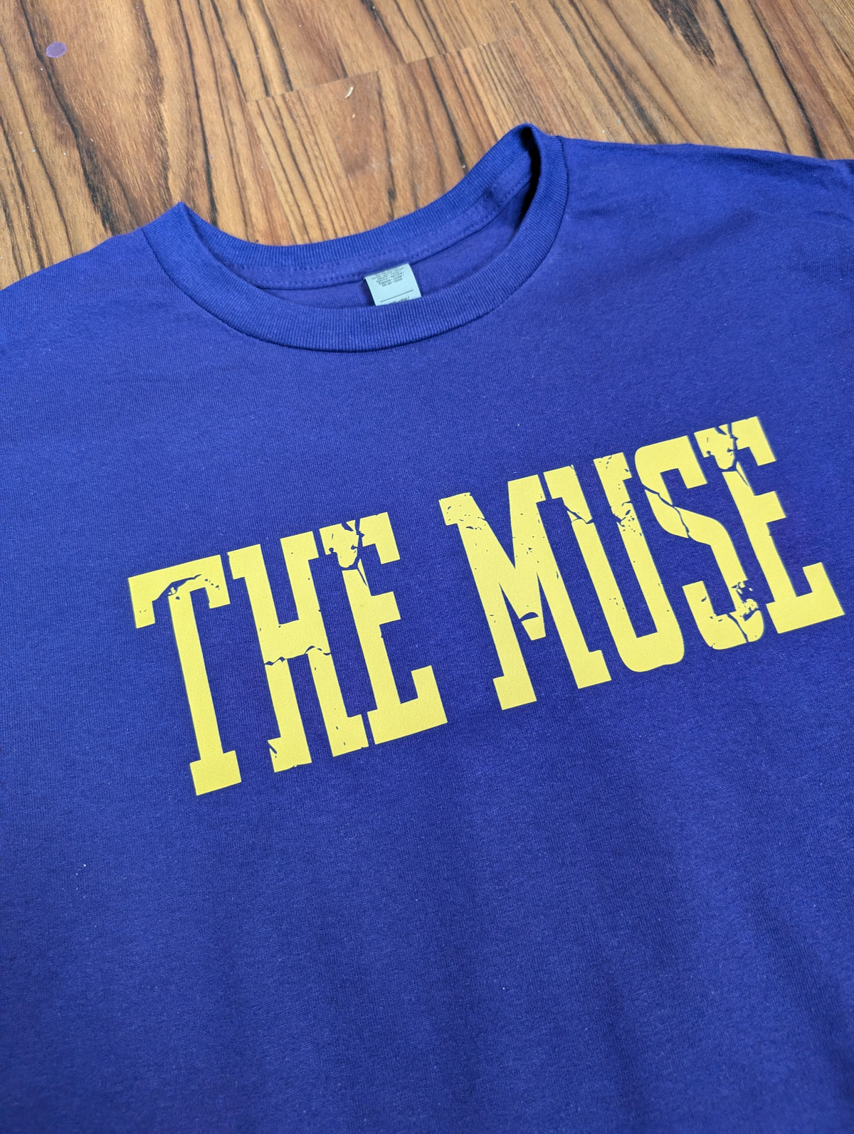 purple t-shirt with 'THE MUSE' text in yellow on a wooden surface