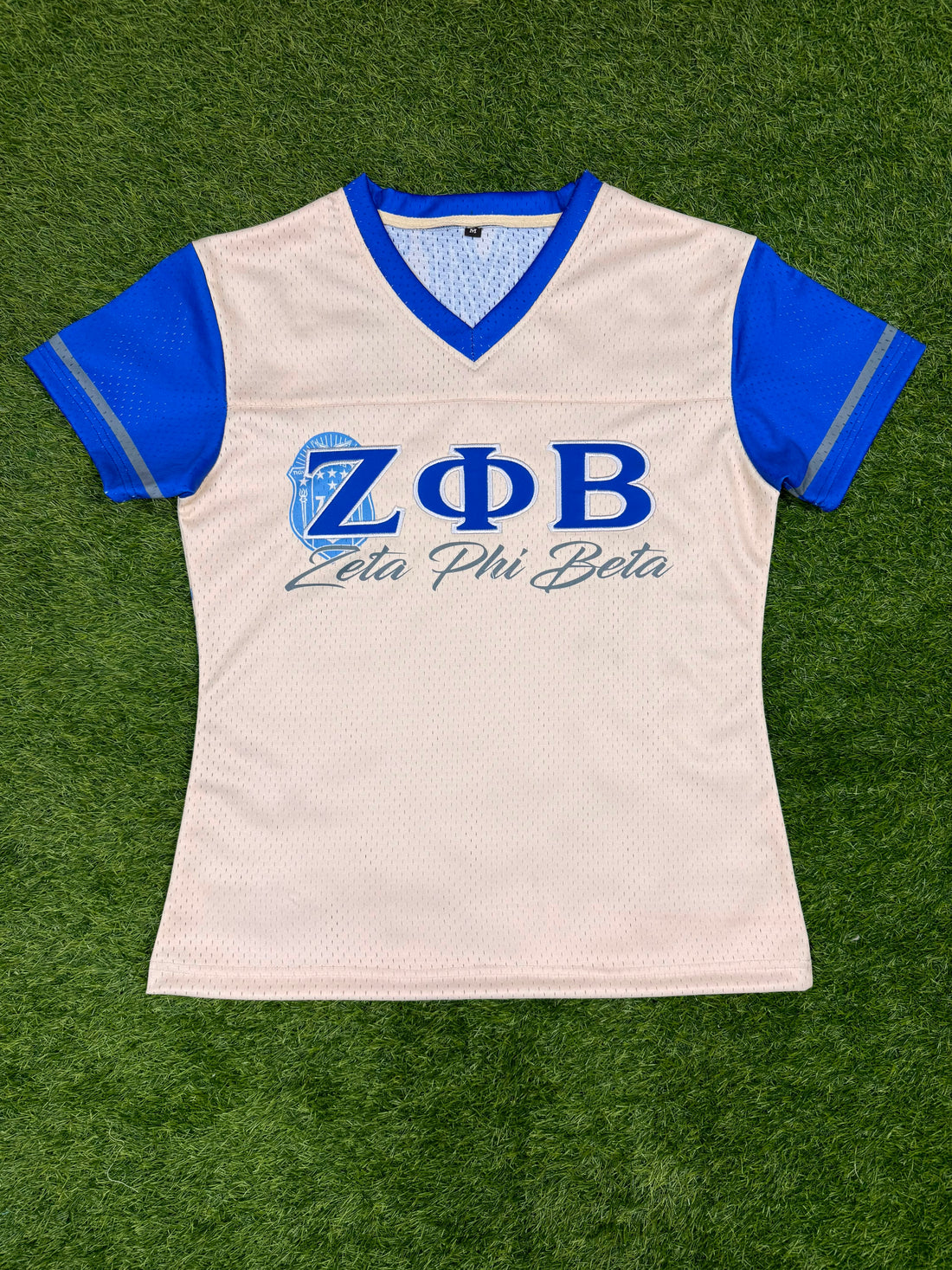 A women's short-sleeve mesh athletic jersey in off-white with royal blue v-neck trim and blue sleeves, featuring the Greek letters Zeta Phi Beta and 'Zeta Phi Beta' printed on the chest. The jersey is laid flat on green artificial turf.