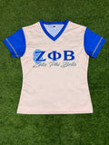 A women's short-sleeve mesh athletic jersey in off-white with royal blue v-neck trim and blue sleeves, featuring the Greek letters Zeta Phi Beta and 'Zeta Phi Beta' printed on the chest. The jersey is laid flat on green artificial turf.