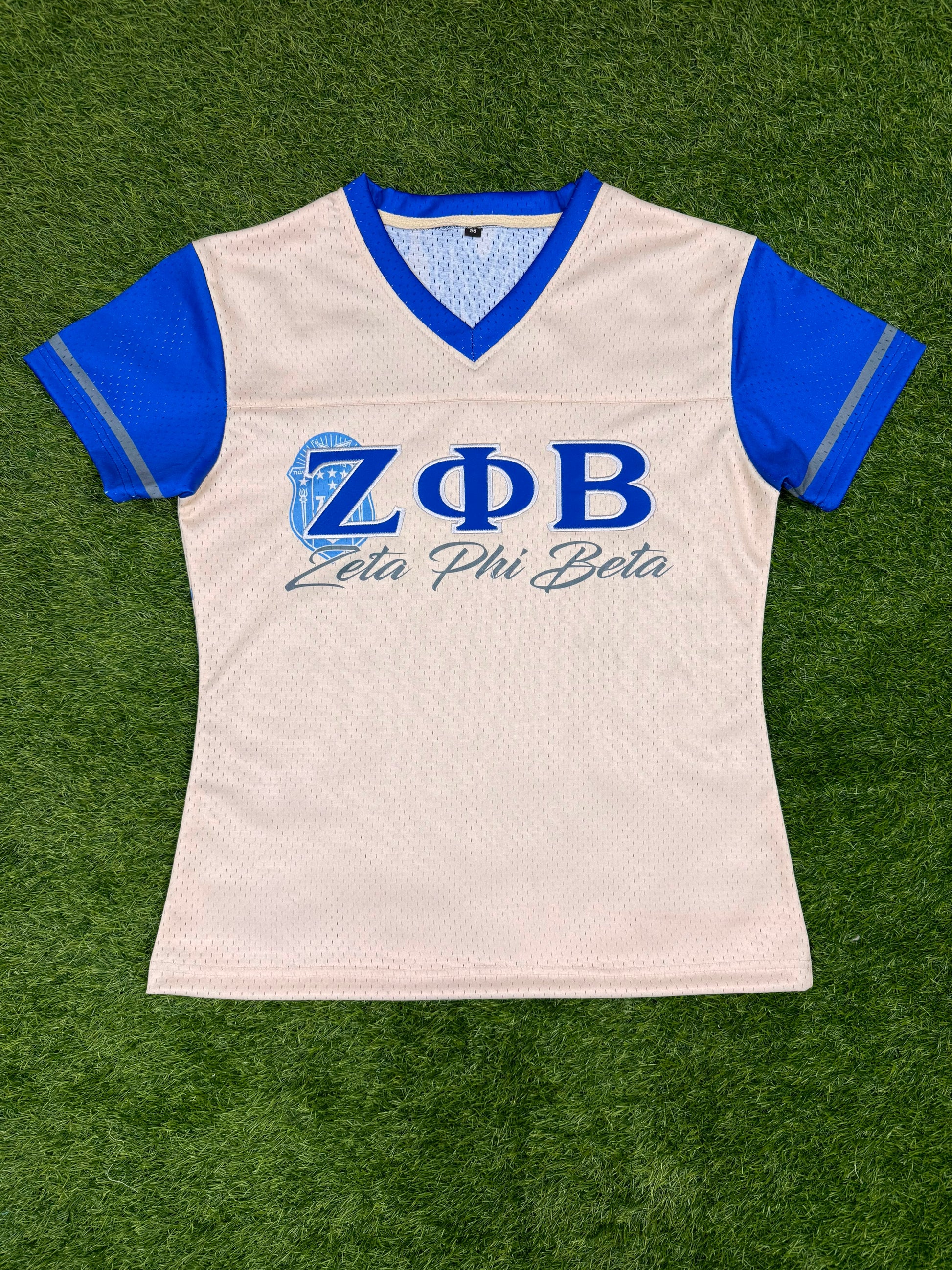 A women's short-sleeve mesh athletic jersey in off-white with royal blue v-neck trim and blue sleeves, featuring the Greek letters Zeta Phi Beta and 'Zeta Phi Beta' printed on the chest. The jersey is laid flat on green artificial turf.