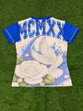 A short-sleeve graphic jersey laid flat on green artificial turf. The jersey has an off white body with blue sleeves and a blue 'MCMXX' print across the upper back, featuring a large white rose and flying white dove graphic on the lower back.