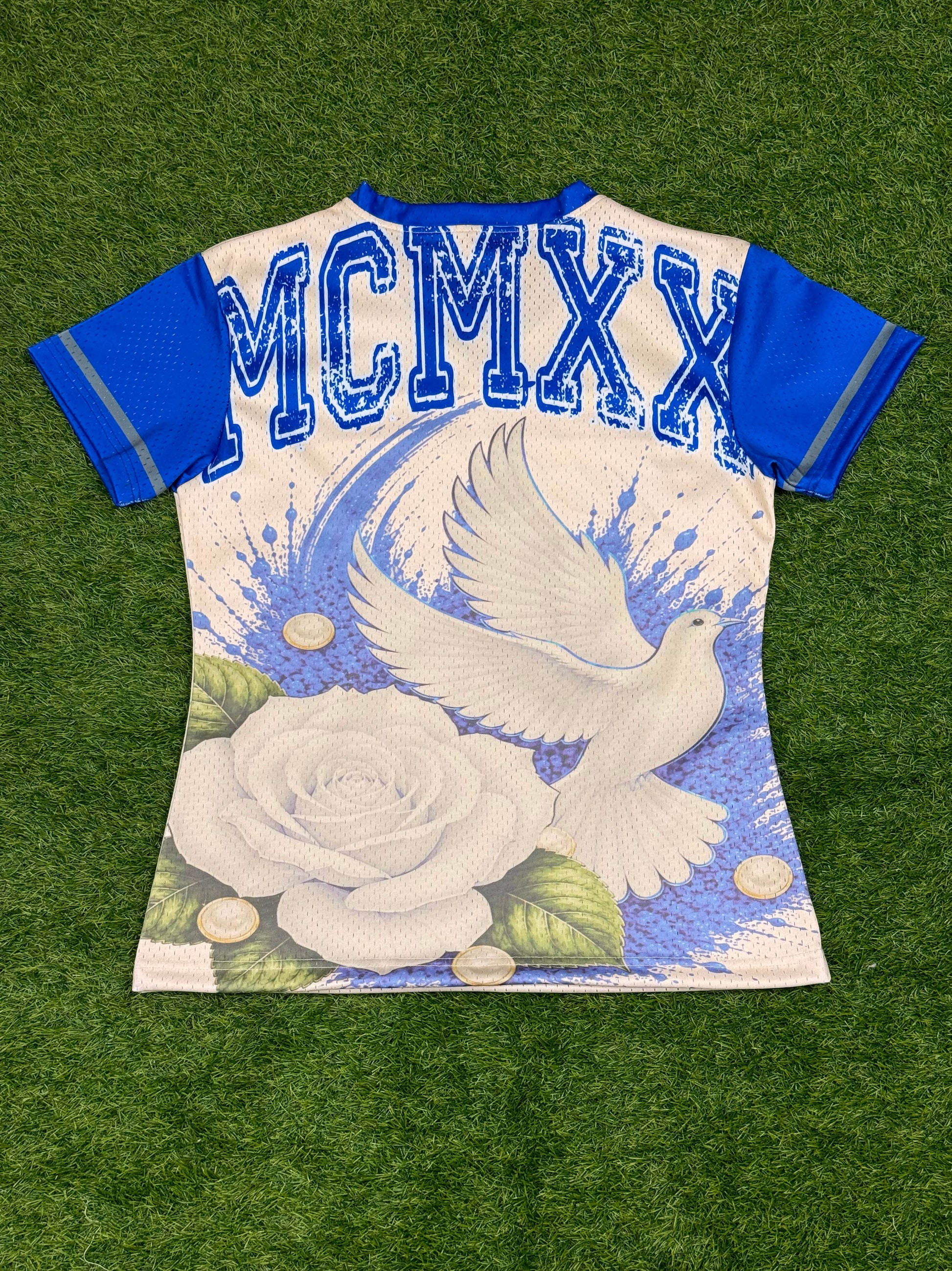 A short-sleeve graphic jersey laid flat on green artificial turf. The jersey has an off white body with blue sleeves and a blue 'MCMXX' print across the upper back, featuring a large white rose and flying white dove graphic on the lower back.