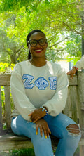 Person wearing a white sweatshirt with blue letters, sitting outdoors under green trees.